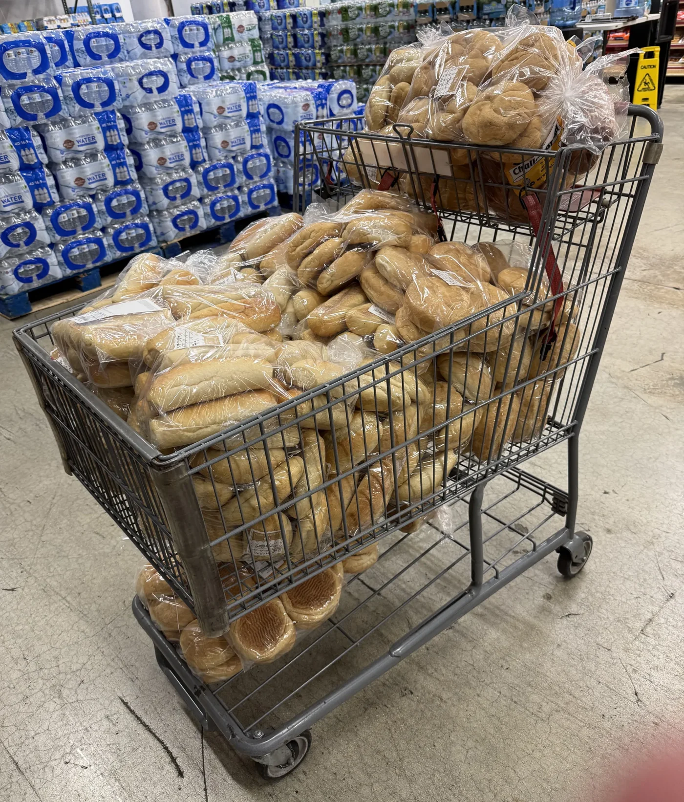 A high-angle shot of a metal shopping cart overflowing with dozens of large bags of sandwich rolls and bread in a warehouse setting, prepped for the NYC Deli theme camp at Love Burn 2026. Stacks of bottled water are visible in the background, illustrating the massive scale of food logistics for the regional Burning Man event.