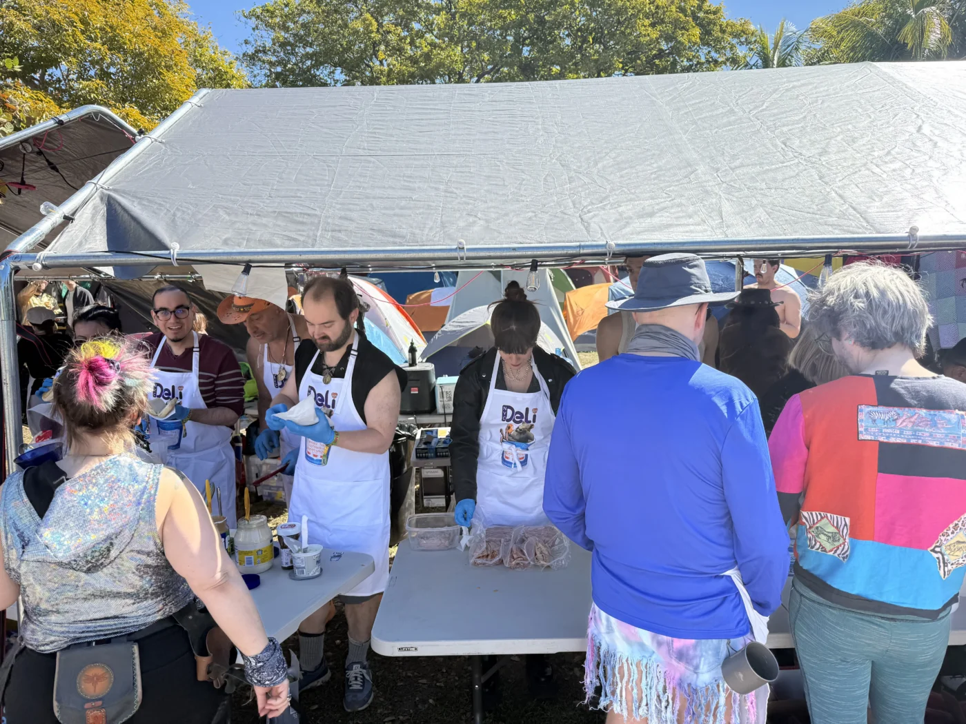 Staff at Camp NYC Deli serving sandwiches under a canopy at an outdoor event, representing hospitality industry community and teamwork.