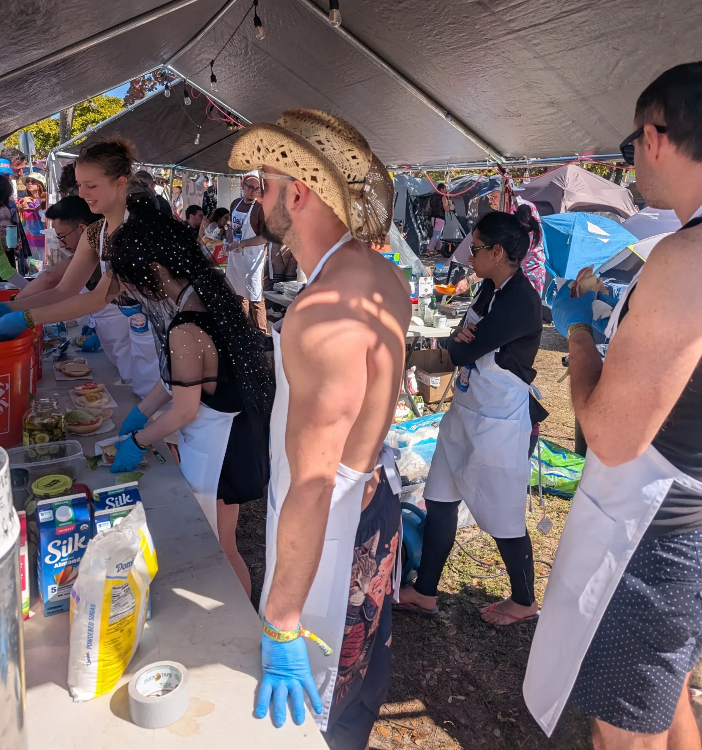 A candid, high-angle shot of the NYC Deli theme camp crew at Love Burn 2026. Staff members wearing white aprons and blue latex gloves work behind a long prep table under a tent, assembling sandwiches with various ingredients like pickles, lettuce, and Silk almond milk visible. A man in a straw cowboy hat stands in the foreground, overlooking the busy service line in the sunny outdoor camping area.