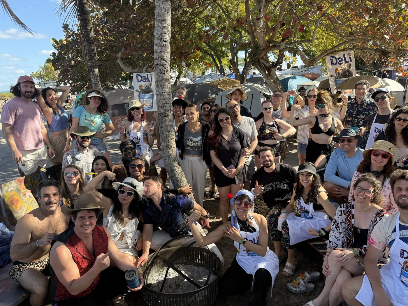 A group photo of the NYC Deli camp staff at a regional Burning Man event, featuring dozens of volunteers in eclectic desert-wear and aprons posing under trees.