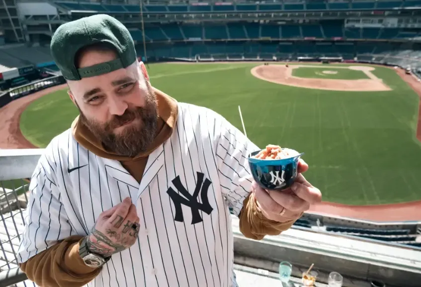 Chef Christian Petroni wearing a New York Yankees jersey and hat, holding a food sample at Yankee Stadium