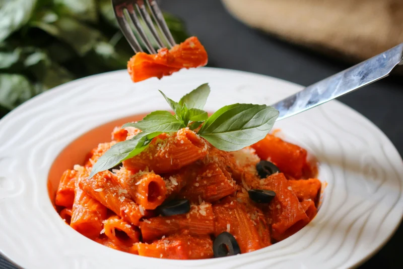 A close up shot of a plate of rigatoni pasta with red tomato sauce, fresh basil leaves, grated parmesan cheese, and black olives