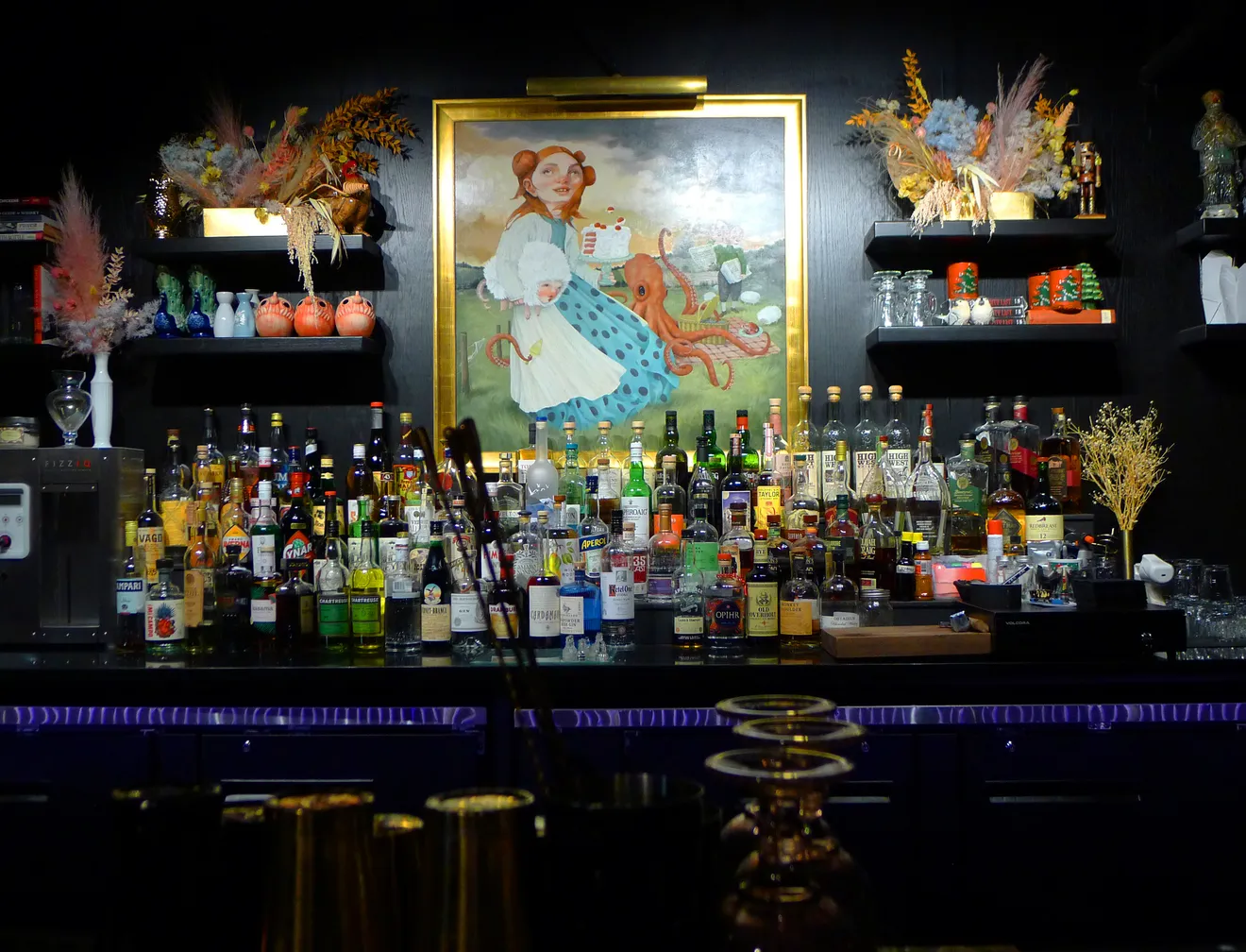A dimly lit, moody cocktail bar backbar stocked with premium liquor bottles, featuring a large surrealist oil painting of a girl with an octopus under a spotlight.