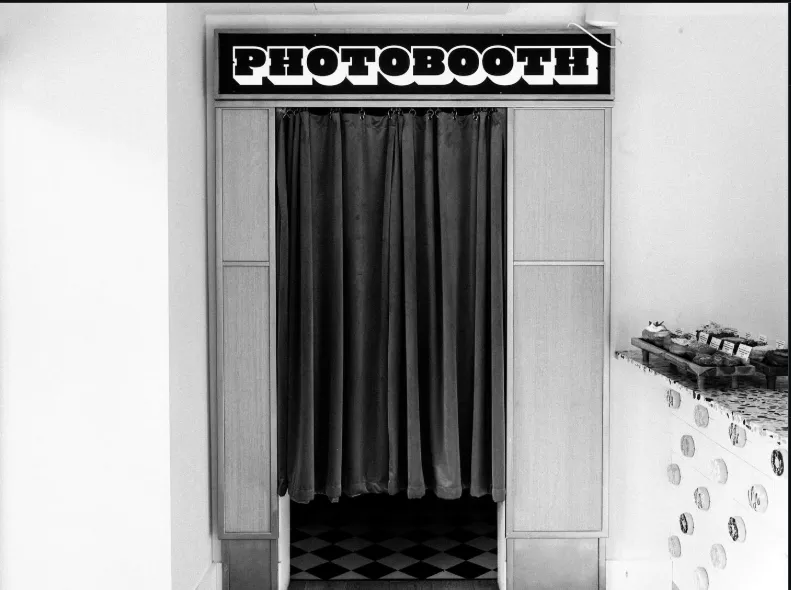 A black and white photograph inside a doughnut shop showing a vintage wood-paneled photobooth with a dark curtain and a checkered floor, with a tray of frosted doughnuts visible on a counter to the right