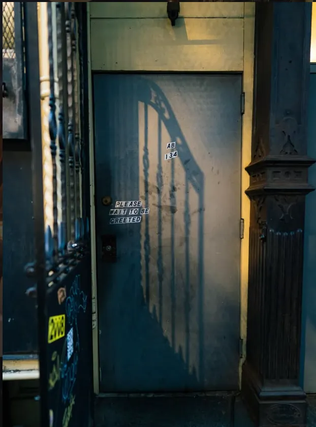 A gray unmarked industrial metal door on the Lower East Side with a sticker that reads 'Please Wait To Be Greeted' and small lettering 'AB 134' under a shadow cast by an iron gate