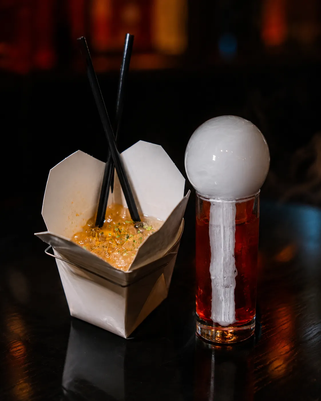 A side-by-side display of two craft cocktails from Good News Darling: one served in a white takeout container with black chopstick straws, and the other a red cocktail in a tall glass topped with a white vapor bubble and white paint on the glass.
