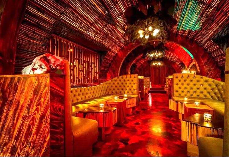 The interior of a dimly lit speakeasy with an arched ceiling lined with bamboo sticks, plush tufted booths, glowing candlelight, and a fiery red and yellow patterned floor.