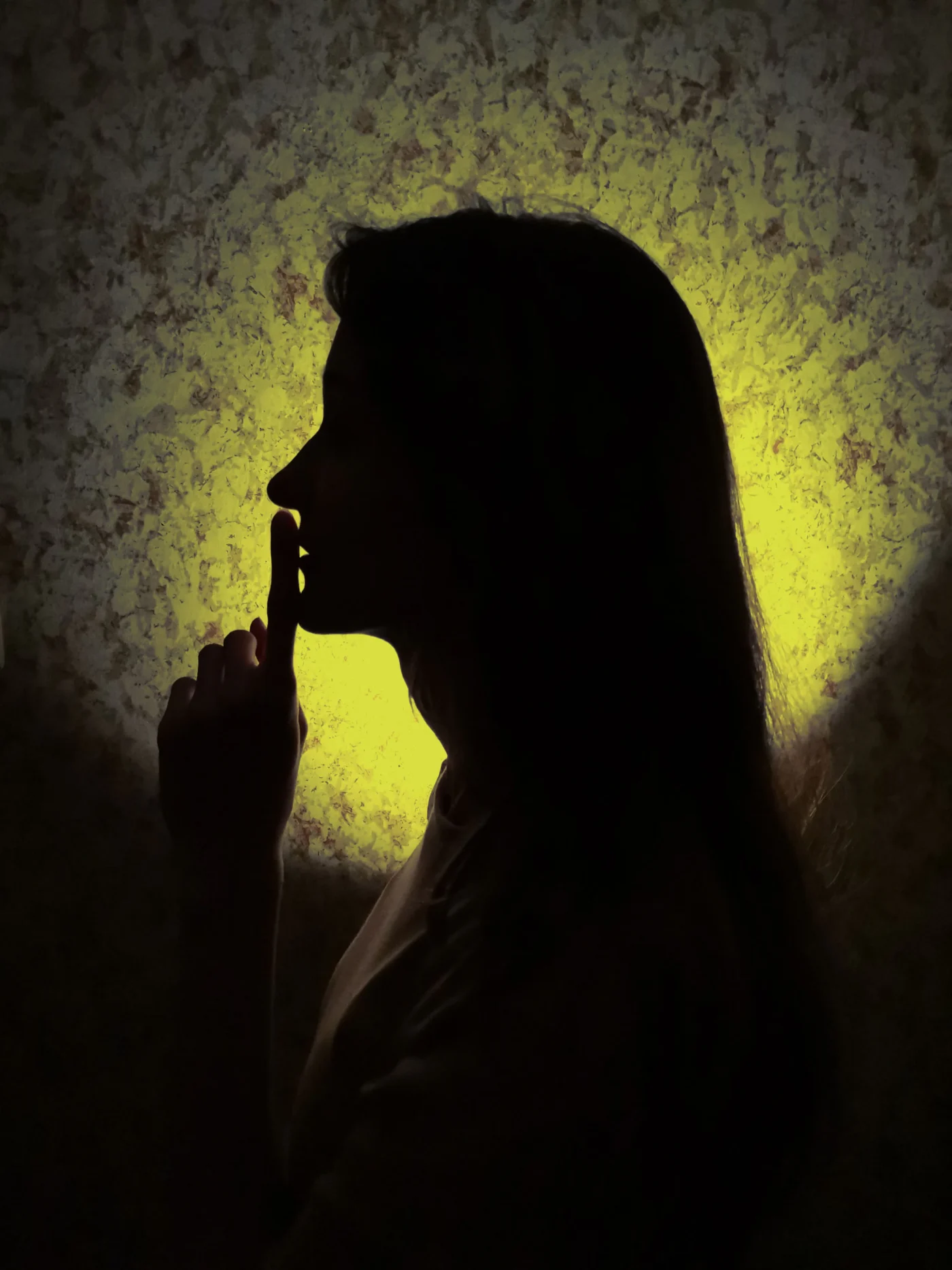A side profile silhouette of a woman holding a finger to her lips in a shushing gesture, backlit by a moody circular yellow glow against a textured wall.