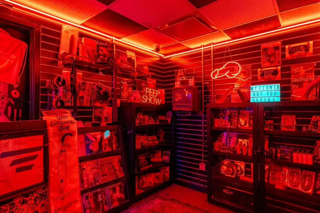 A dimly lit room styled as a 1980s adult novelty shop drenched in intense neon red lighting, featuring shelves stocked with vintage magazines, a retro tube television, and a neon sign