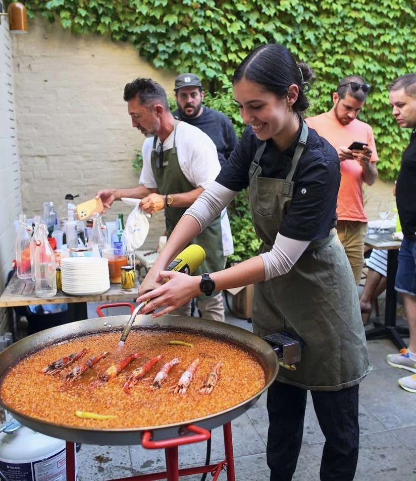 Chef Silvia Garcia-Nevado using a kitchen torch on a giant pan of paella topped with red prawns at an outdoor event.