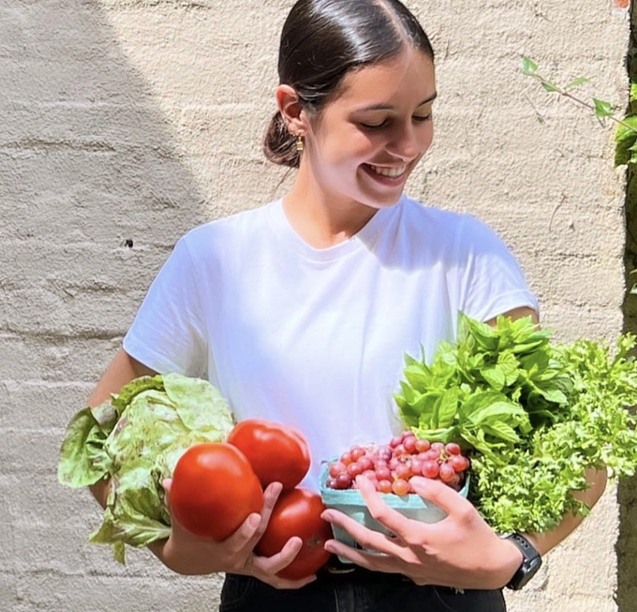 Chef Silvia Garcia-Nevado holding armfuls of fresh produce including heirloom tomatoes, lettuce, grapes, and fresh herbs.