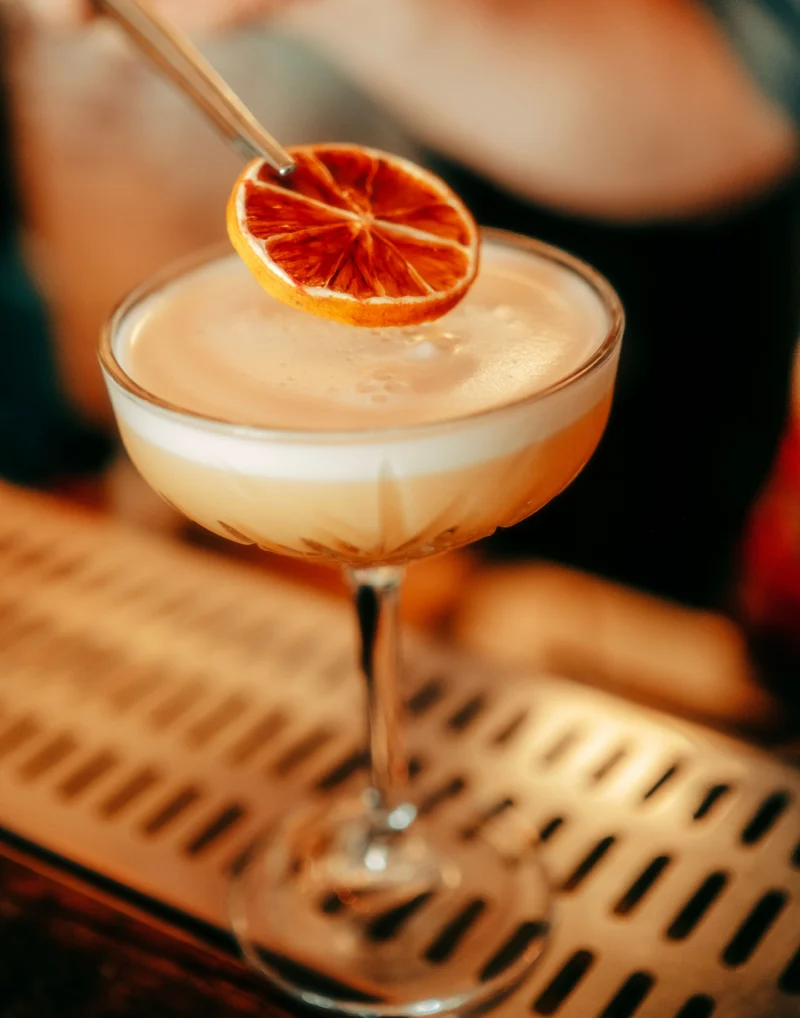 A bartender garnishing a foamy craft cocktail with a dehydrated citrus wheel in a coupe glass for Last Call Cocktails and Shots Part II crossword puzzle